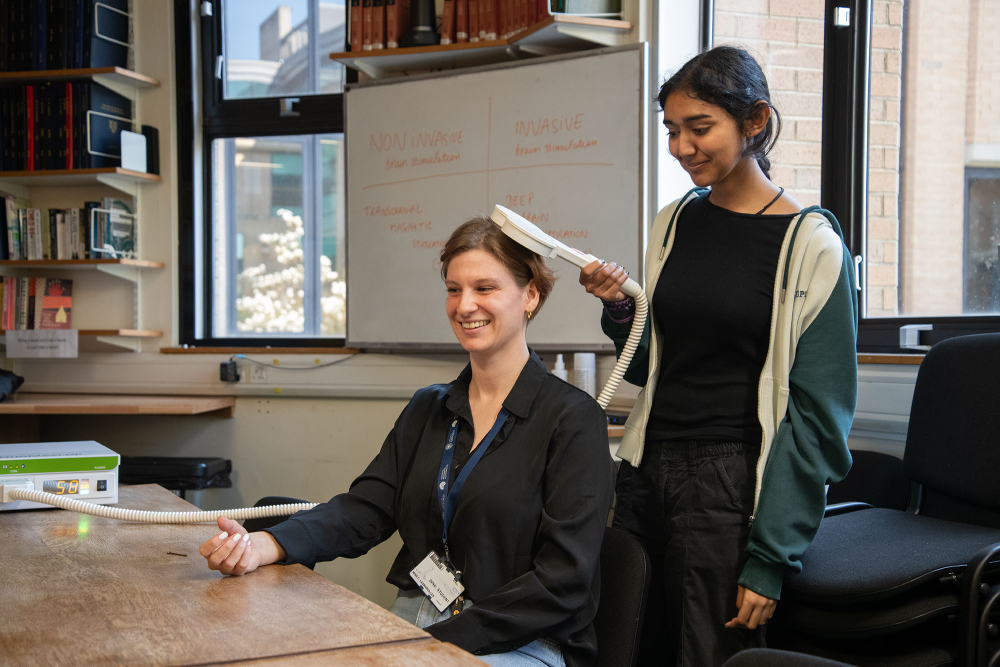a researcher (seated) undergoes transcranial magnetic stimulation, directed by a pupil holding the stimulation coil over their head